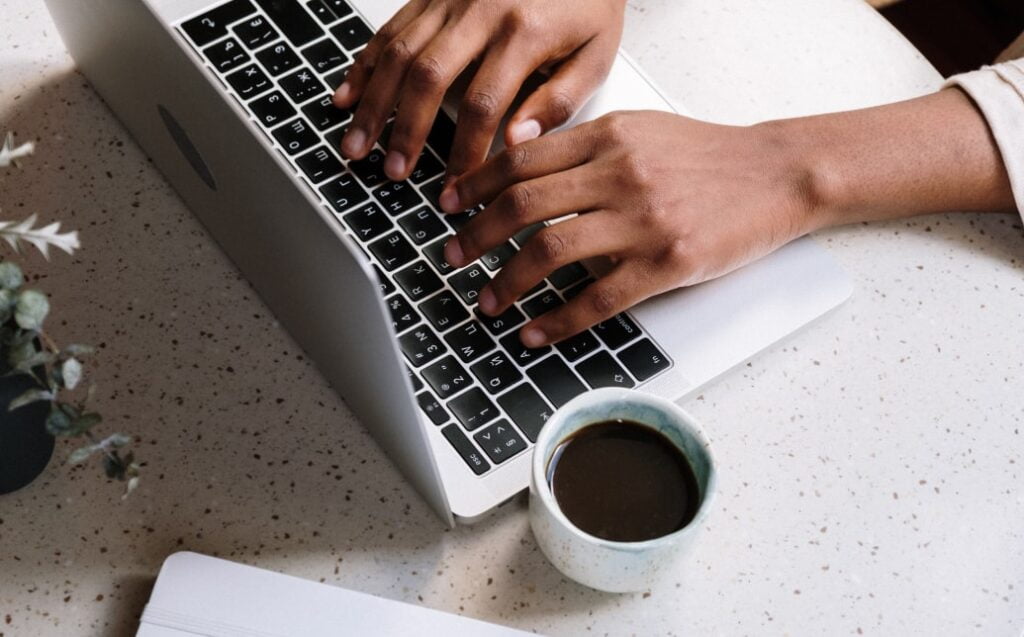 Osoba píšící na notebooku s šálkem kávy na pracovním stole.